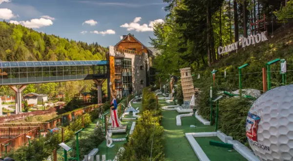 Mini golf z atrakcjami, budynek i las w tle.
